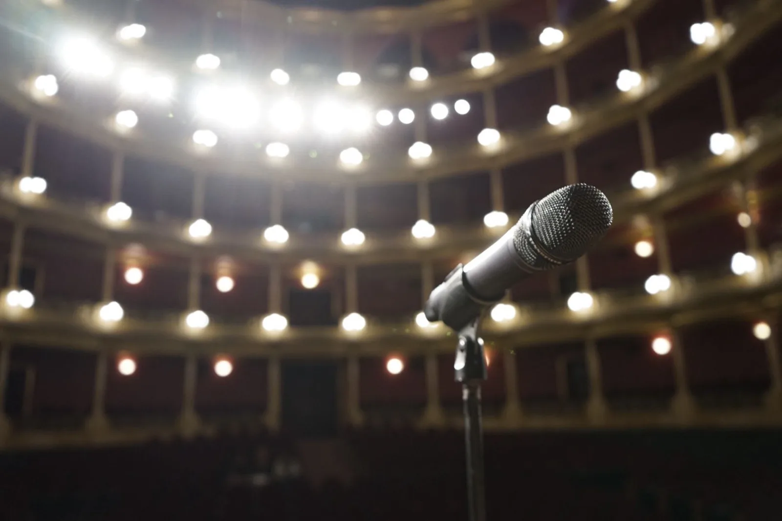 A microphone on a stand, lit by the out-of-focus house lights of an empty venue.